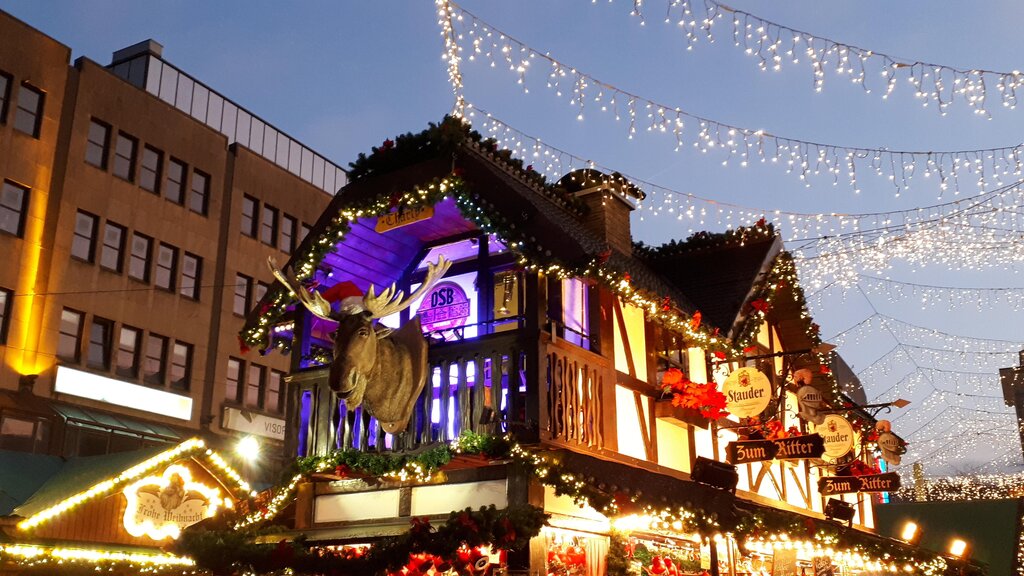 A cozy wooden chalet-style stall at a German Christmas market glows with warm lights, topped with a large moose head decoration and surrounded by festive garlands and twinkling string lights overhead.