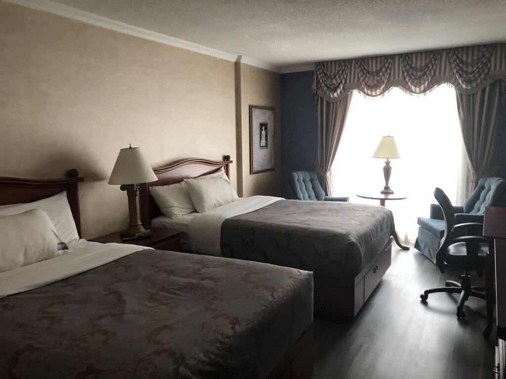 A hotel room in Quebec city with two double beds features classic wooden headboards, matching bedside lamps, and a window dressed with heavy drapes, letting in soft natural light over a seating area with two armchairs and a small table.