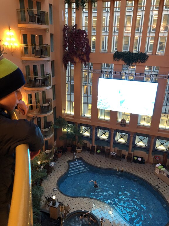 A child looks down from an upper floor balcony at an indoor pool below, set in a tall atrium-style hotel in Quebec City with potted plants, balconies, and a large screen mounted on the wall above the water.