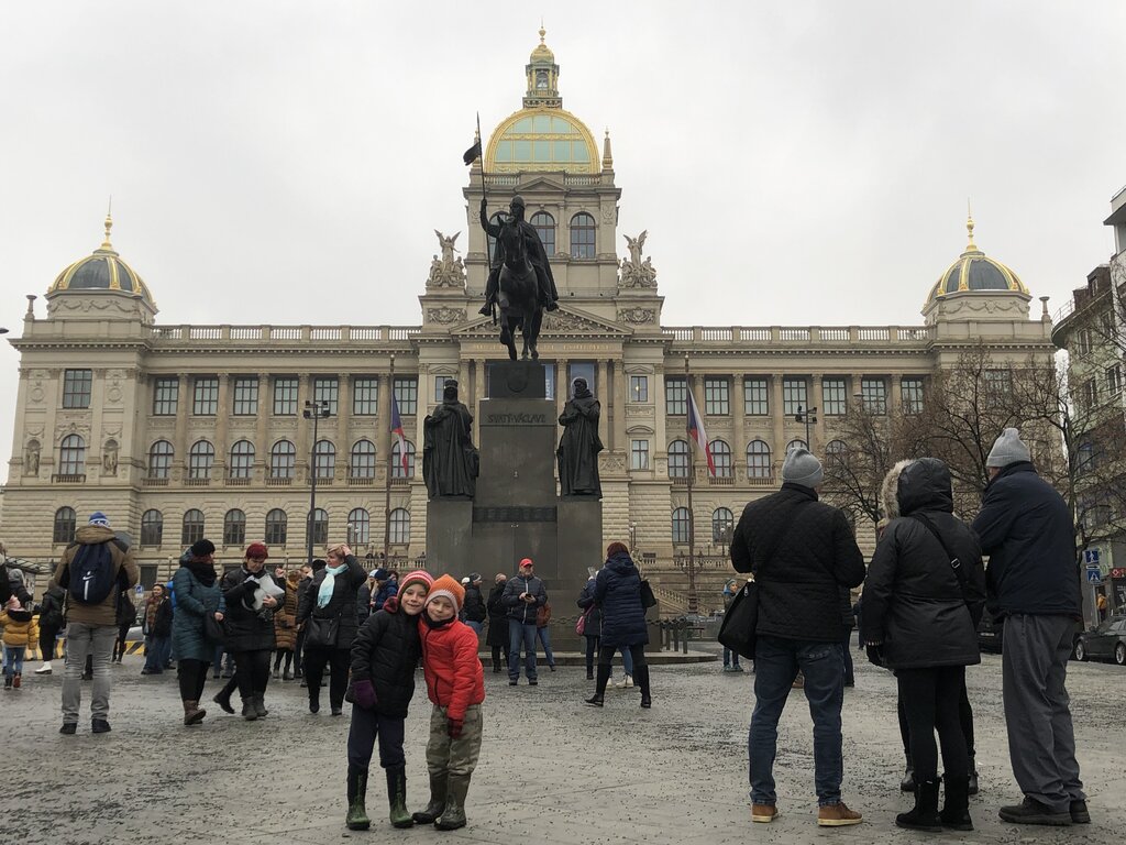 Two young children in winter clothes pose and smile in front of a large equestrian statue of Saint Wenceslas at Wenceslas Square in Prague. Behind them stands the grand façade of the National Museum, with its ornate domes and Czech flags, as crowds of people gather around the plaza on a gray, overcast day.