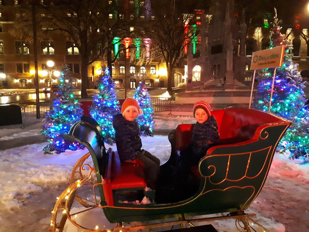 Two children dressed in winter jackets and hats sit in a festive green and red sleigh surrounded by lit Christmas trees in a snowy city square at night, with colourful lights and a Desjardins sign in the background.