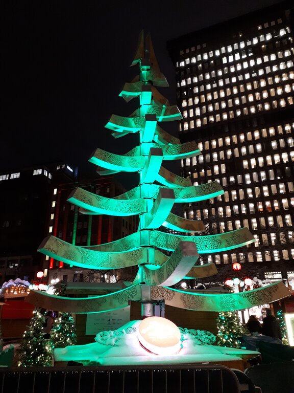 A modern, stylized Christmas tree sculpture glows green in the centre of a holiday market at night, with illuminated buildings and decorated evergreens surrounding it.