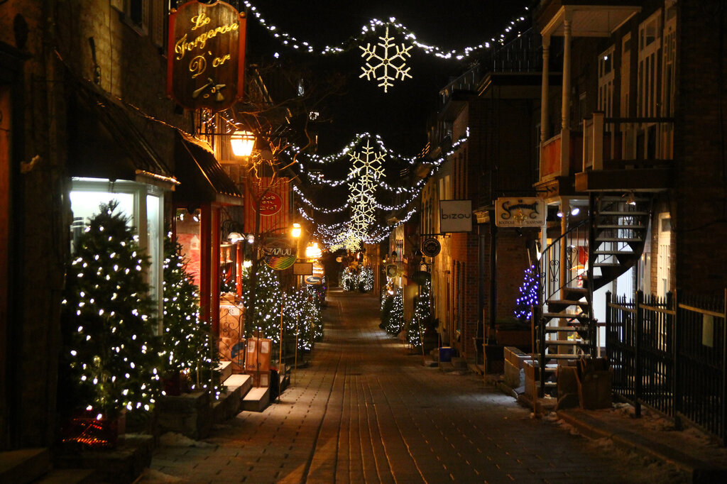 A charming pedestrian street in Petit Champlain Quebec City is lined with lit Christmas trees and hanging snowflake lights stretches between boutique shops, creating a cozy and festive nighttime atmosphere.