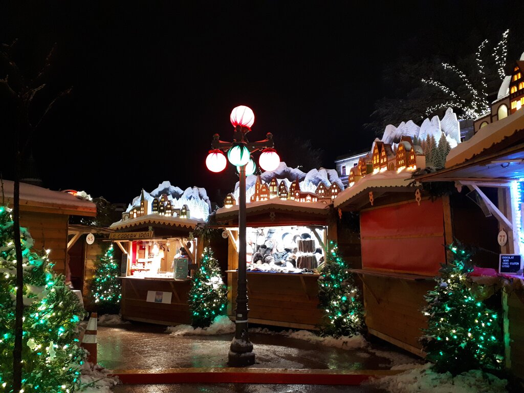 Christmas market stalls in Quebec City are decorated with snow-covered miniature village rooftops and strings of lights are set up around a red and green lamppost, with shoppers browsing in the snowy scene.
