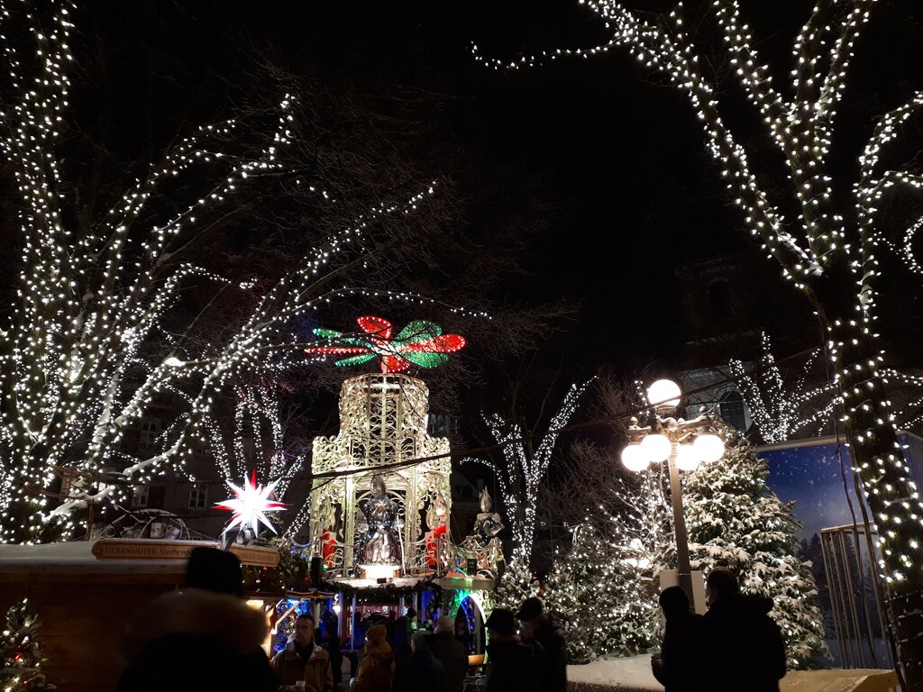 Twinkling white lights wrap around bare trees in a festive night scene where people gather around an ornate Christmas pyramid with spinning red and green blades and a nativity display below.
