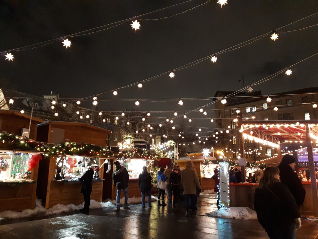 A night scene at the Quebec City Christmas market with wooden booths lit by festive garlands, and glowing star-shaped lights strung overhead as people shop and stroll on the wet pavement.