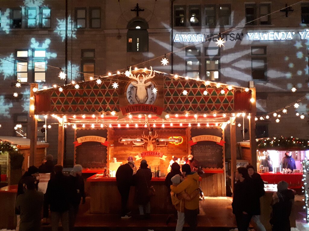 A wooden Winterbar booth decorated with warm string lights and star ornaments serves drinks to visitors at the outdoor QUebec City German Christmas market, with snowflake projections on the stone building behind.