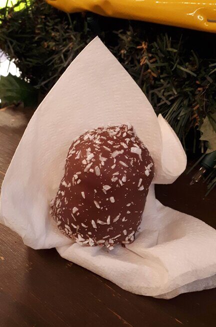 A chocolate-coated marshmallow treat sprinkled with white coconut flakes is nestled in a folded napkin, sitting on a wooden counter with holiday greenery in the background.