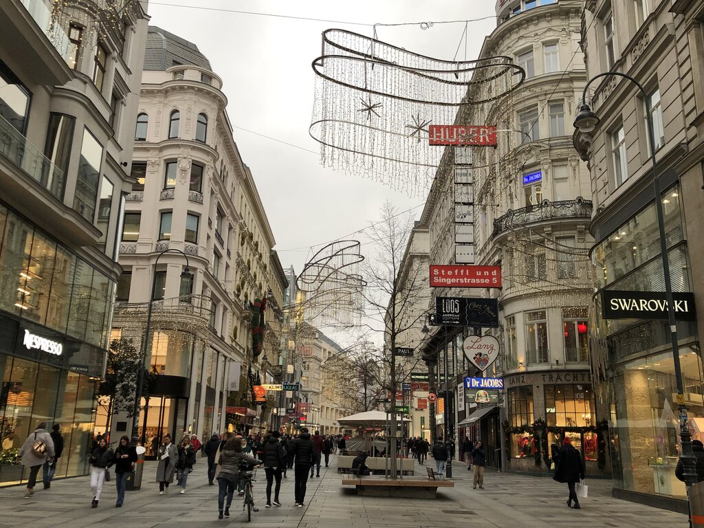 Busy pedestrian shopping street in Vienna decorated with festive hanging lights and holiday displays. Shoppers walk past stores like Swarovski and Nespresso, surrounded by ornate historic buildings.