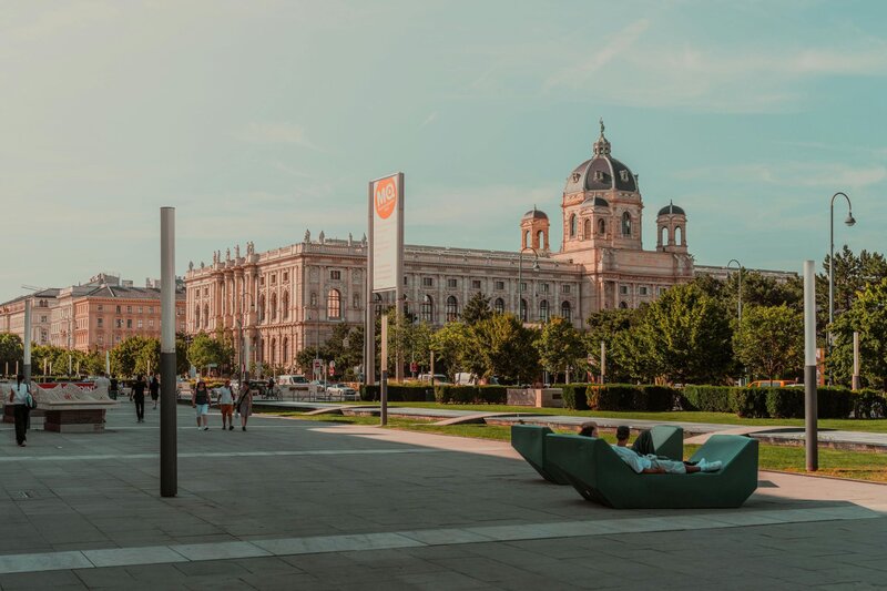 People relax on modern outdoor seating in Vienna’s MuseumQuartier, with the grand dome and symmetrical towers of the Kunsthistorisches Museum rising behind a line of trees. The plaza features clean lines, art installations, and a bright afternoon sky.