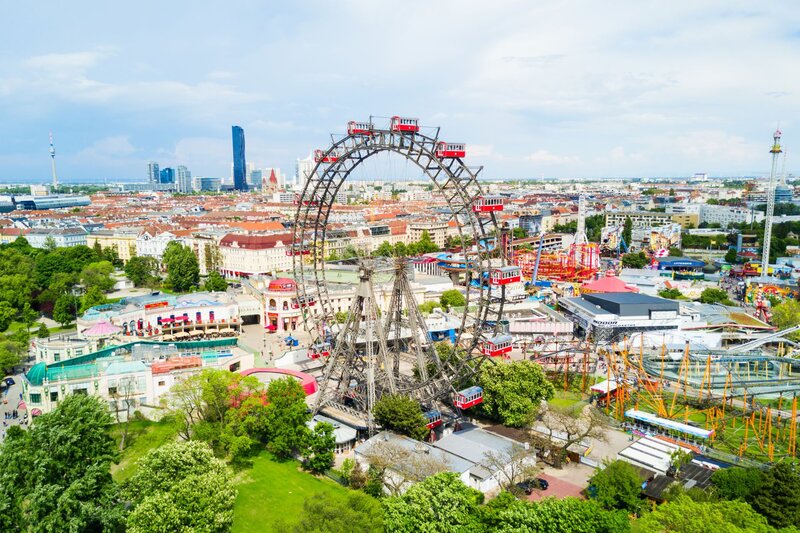 Aerial view of Vienna's Prater amusement park featuring the iconic Wiener Riesenrad, a large Ferris wheel with red gondolas. The colourful park rides and city skyline stretch out into the distance on a bright day.