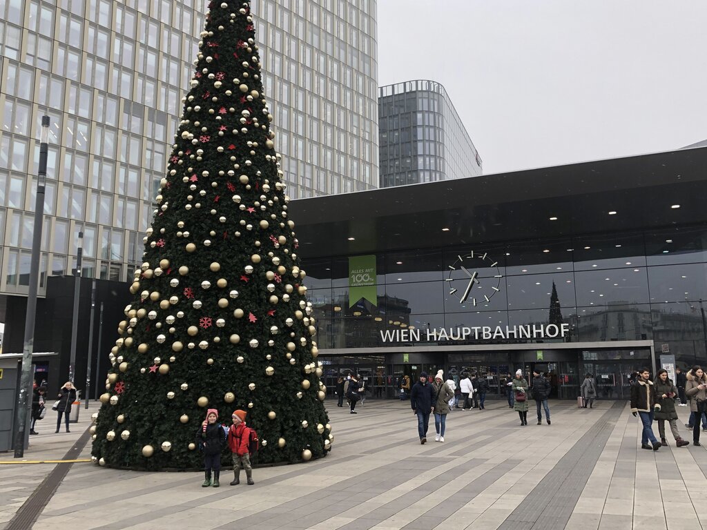 Tall Christmas tree decorated with red and gold ornaments stands in front of Wien Hauptbahnhof, Vienna’s main train station. People walk by and two children in winter clothes stand at the base of the tree.