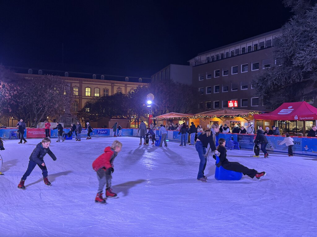 Children and adults skate under twinkling lights at a lively outdoor ice rink near the Trier Christmas market, with festive stalls and sponsor banners in the background.