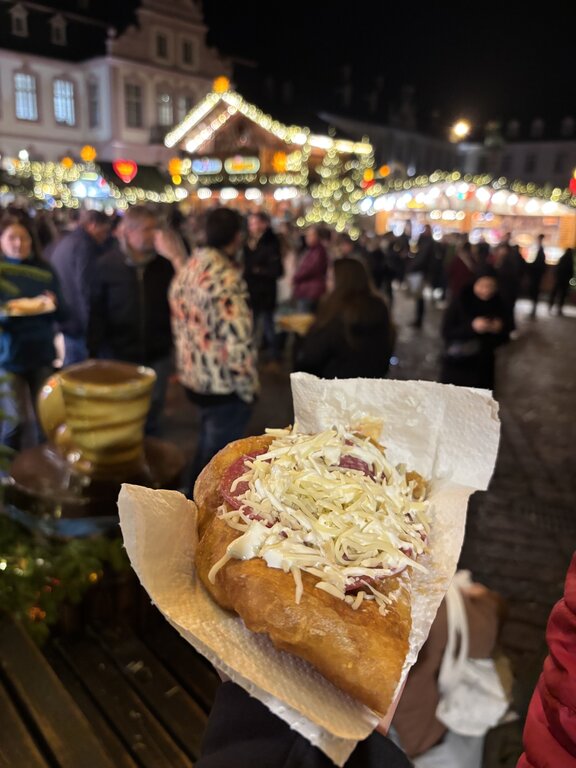 A hand holds a traditional fried bread snack called langos, which is topped with cheese at the Trier Christmas market, with festive lights and shoppers in the background.