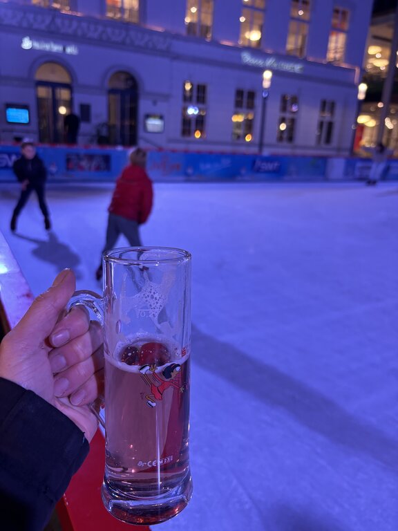 A hand holds a clear glass of hot drink above an outdoor ice rink in Trier, where children skate in the background under soft purple-blue lighting.