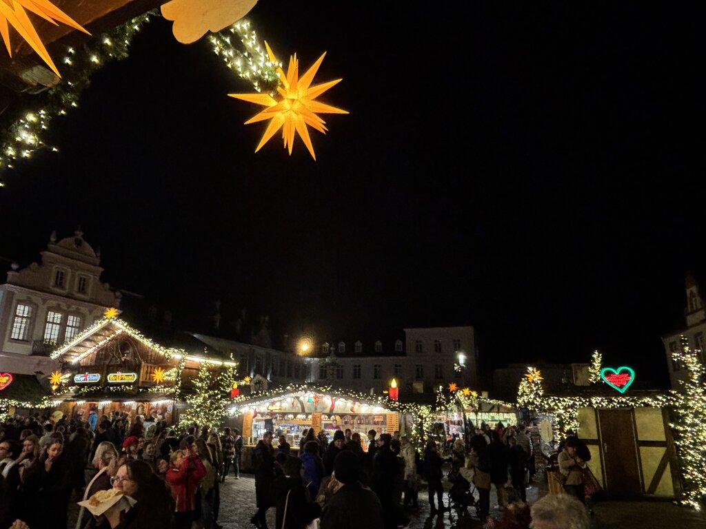Crowds gather at the Trier Christmas market under glowing star lanterns and twinkling lights, with wooden stalls offering food and gifts in the town square.
