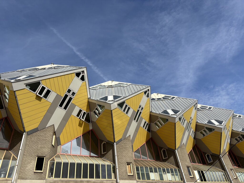 Tilted yellow cube-shaped houses with slanted roofs and geometric windows, set against a bright blue sky. The bold modern architecture stands out with angular lines and contrasting materials.