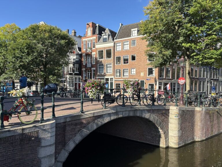 A charming brick canal bridge in Amsterdam covered with parked bikes and flower baskets under sunny skies. A red bike adorned with white flowers leans against the rail near a tree-lined street with classic brick houses.