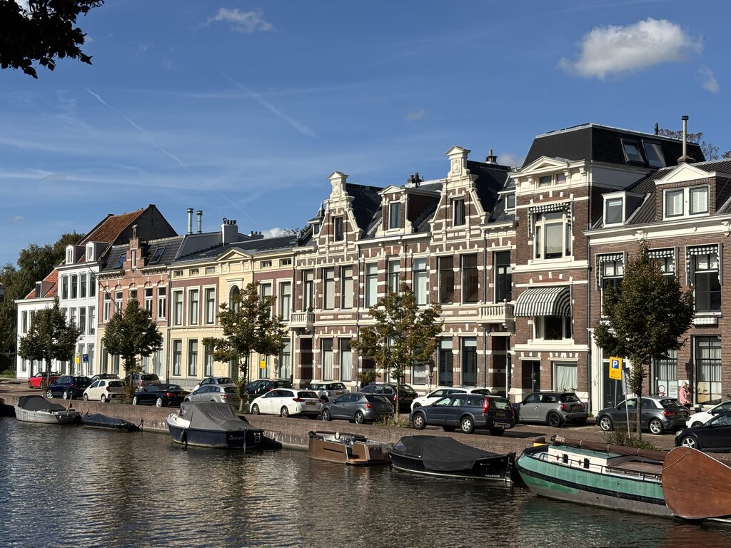Ornate historic townhouses line a canal in Haarlem, with parked cars and small boats docked along the edge. The buildings feature detailed brickwork, pastel colours, and traditional Dutch architectural styles.