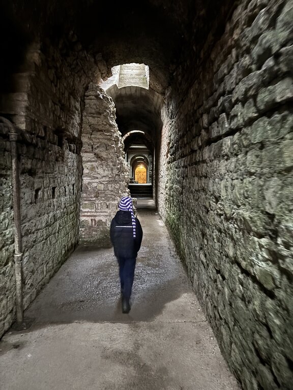 A child in a long striped hat walks through a long, dimly lit stone corridor inside an ancient Roman structure in Trier, surrounded by textured historic walls.