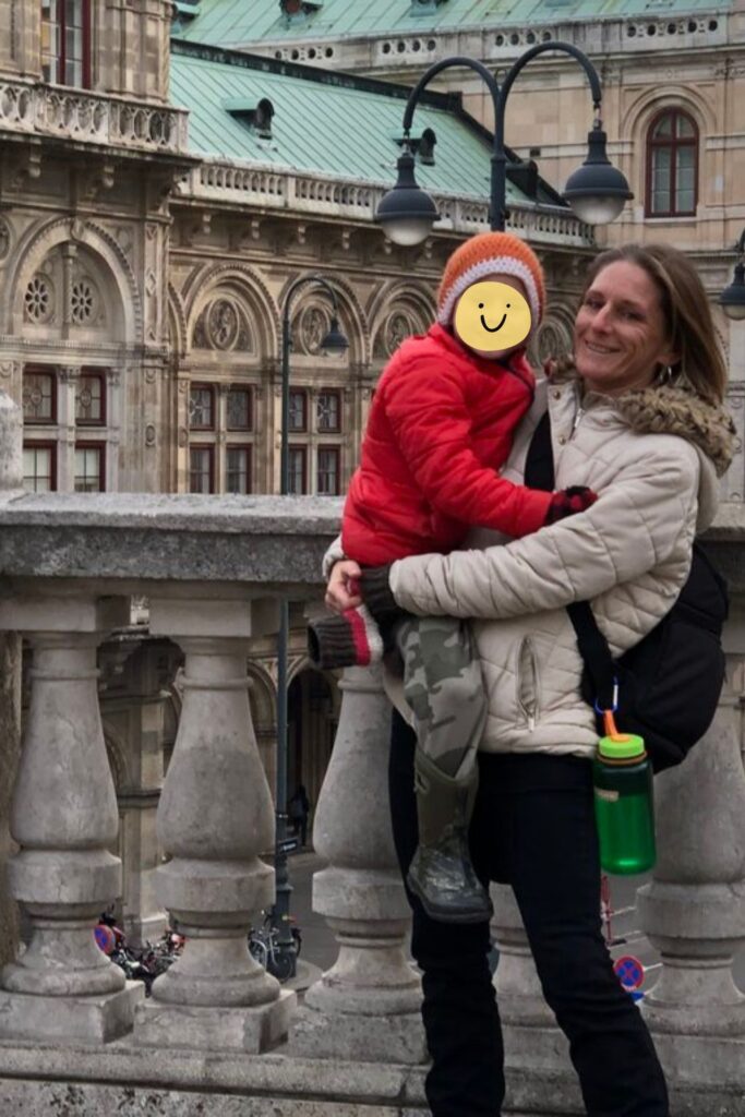 A smiling woman in a beige winter coat holds a child in a red jacket and orange knit hat on a stone balcony, with an ornate historic building and street lamps in the background. The child’s face is covered with a sticker featuring a smiley face.