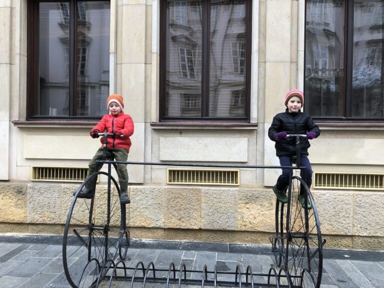 Two young children in winter clothing sit on metal penny-farthing sculptures, posing like they're riding antique bicycles outside a stone building with large windows in a square in Bratislava, Slovakia