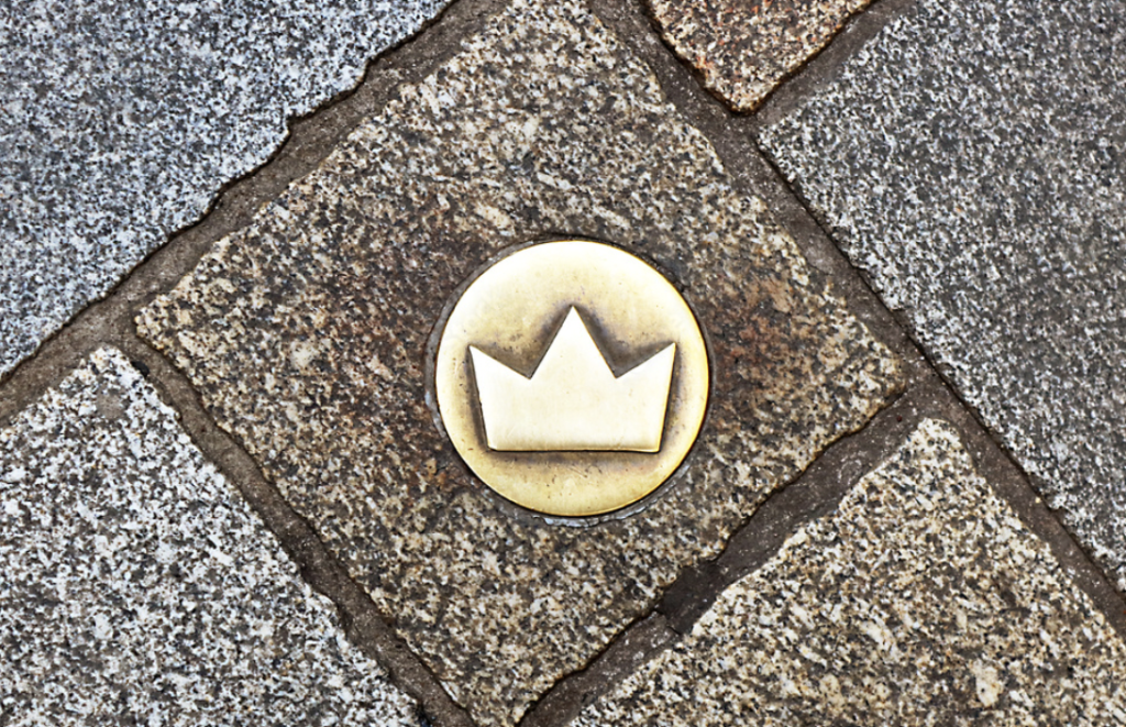 Circular brass marker with a minimalist crown symbol embedded in a cobblestone sidewalk. The surrounding stones form a diagonal pattern in varying shades of grey and brown.