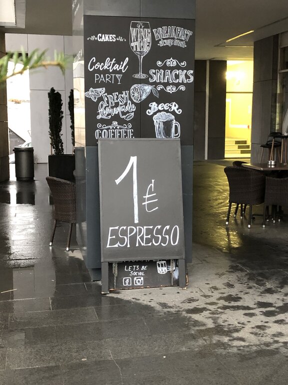 A sidewalk chalkboard sign outside a café advertises espresso for 1 euro, with playful chalk illustrations above it promoting cakes, cocktails, fresh lemonade, coffee, beer, breakfast, and snacks. The seating area nearby features wicker chairs and wet pavement from recent rain.