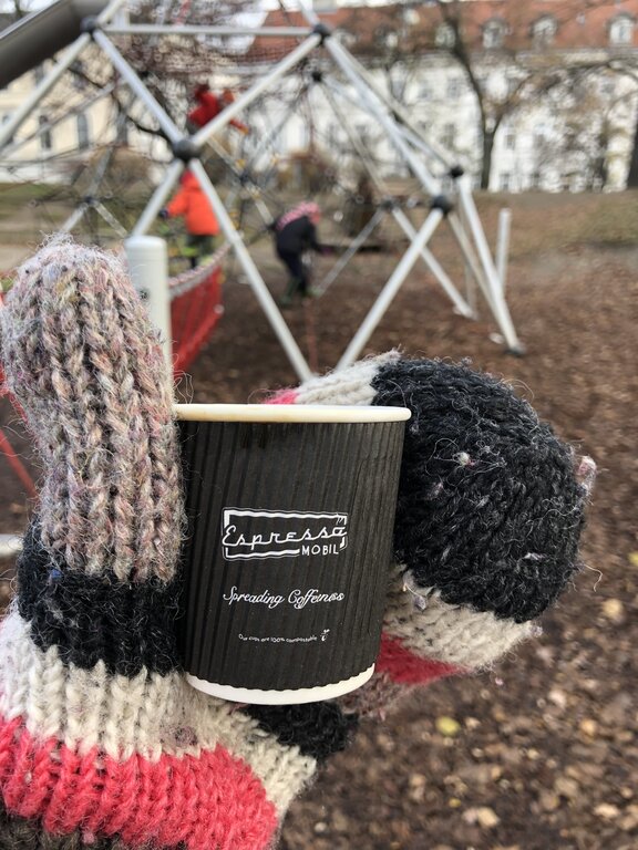 A gloved hand holds an Espresso Mobil coffee cup in the foreground, with children playing on a geometric rope climbing dome at Stadtpark playground in Vienna during winter.