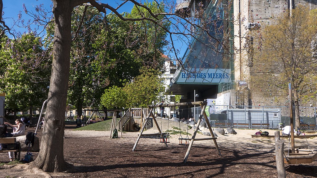 A woodchip-covered playground in Esterházypark, Vienna, with swings and a climbing structure set beneath tall trees, next to the Haus des Meeres urban zoo and climbing wall.