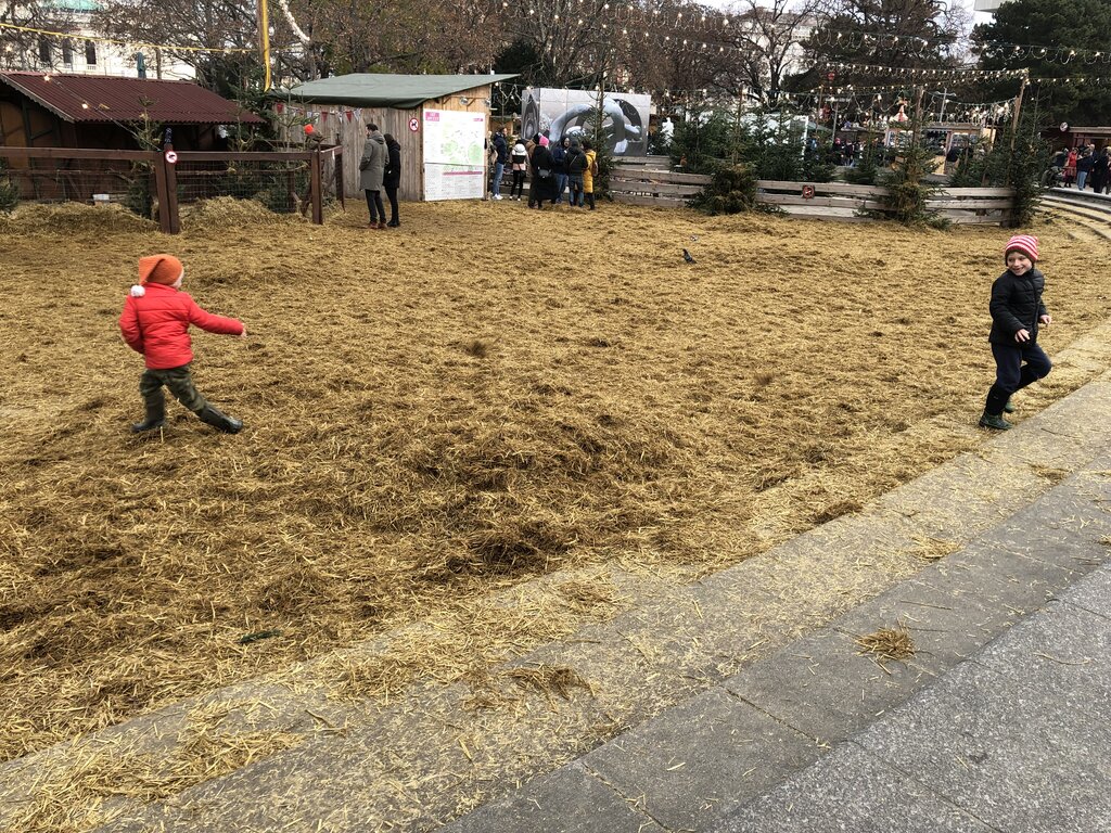 Two children run across a large straw-covered play zone set up for winter festivities in Vienna, with holiday decorations, pine trees, and wooden booths in the background.
