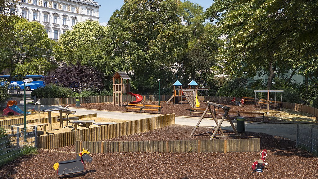 A spacious, fenced playground at Rathausplatz in Vienna, featuring slides, swings, climbing towers, and seating areas, surrounded by tall trees and elegant historic buildings.