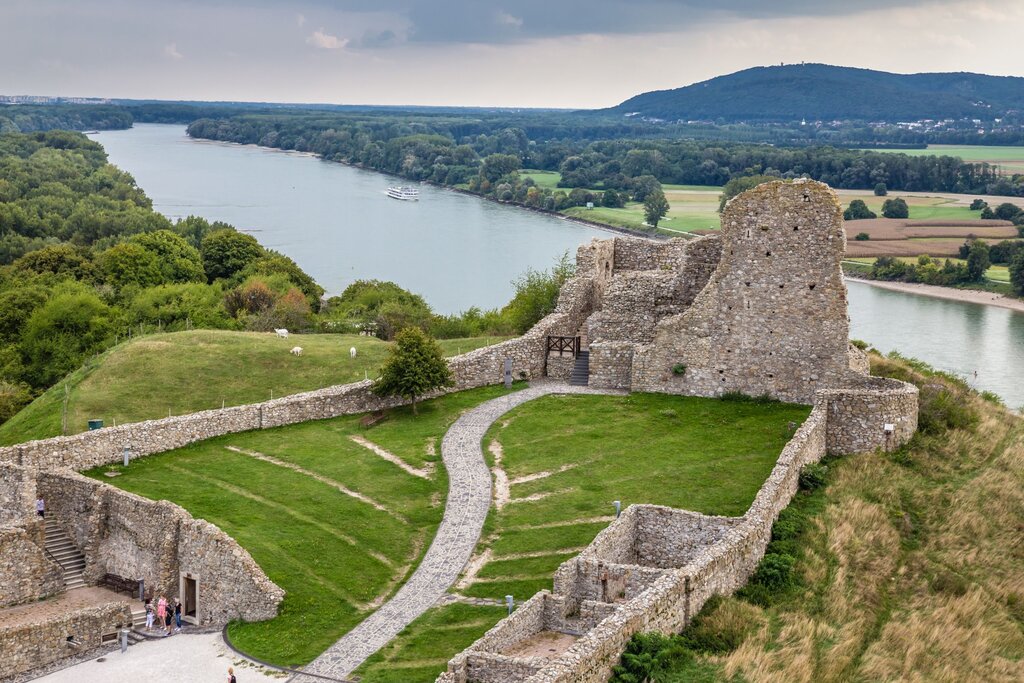Aerial view of Devín Castle ruins perched above the confluence of the Danube and Morava rivers, surrounded by green fields, forest, and a meandering path leading through the stone fortress remains. A riverboat cruises along the Danube under a cloudy sky.