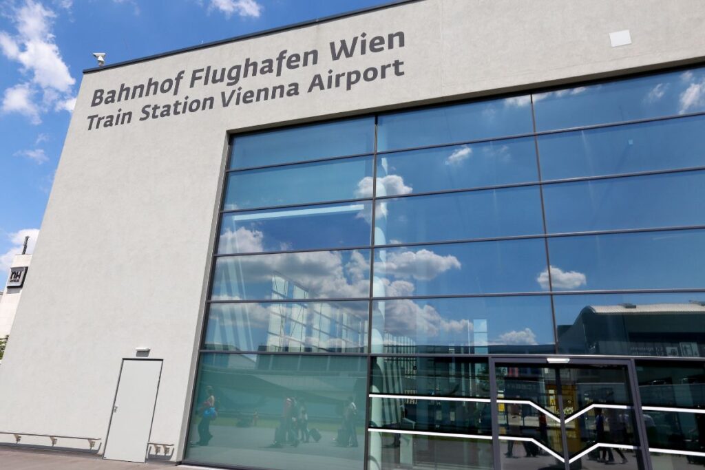 Exterior view of the Bahnhof Flughafen Wien building, with large reflective glass panels mirroring a blue sky with clouds and travellers walking past.