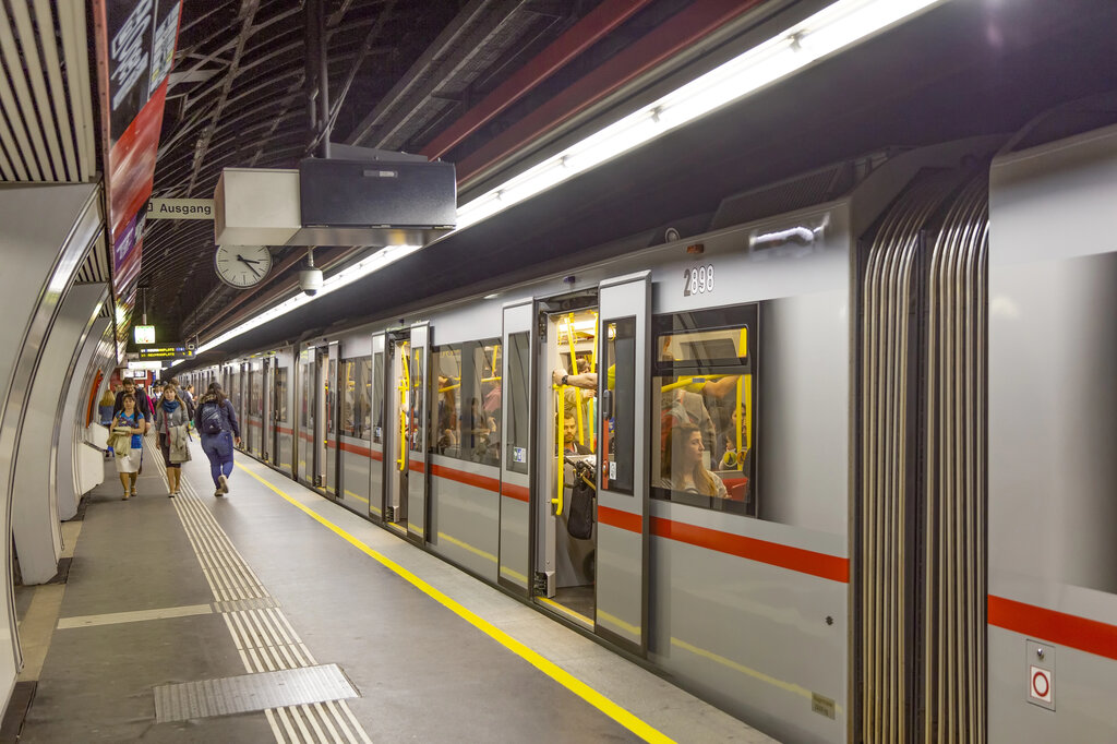 Underground subway platform in Vienna with passengers boarding and exiting a silver and red train under a curved ceiling, with signage and a clock reading "Ausgang" for the exit.