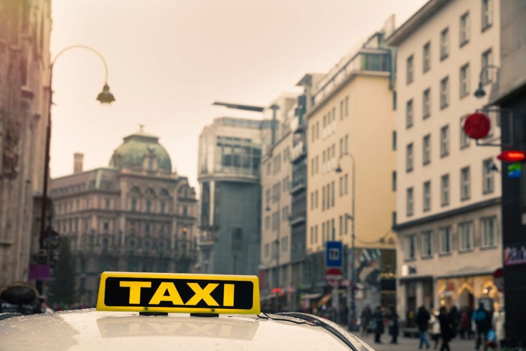 Close-up of a yellow taxi sign on top of a cab in a busy street in Vienna, with historic and modern buildings in the background under a cloudy sky.