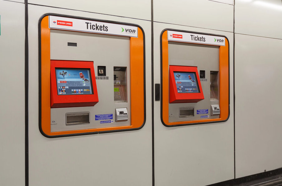 Two orange-framed Wiener Linien and VOR ticket machines with touchscreen interfaces and card slots, mounted on a clean white wall in a Vienna public transit station.