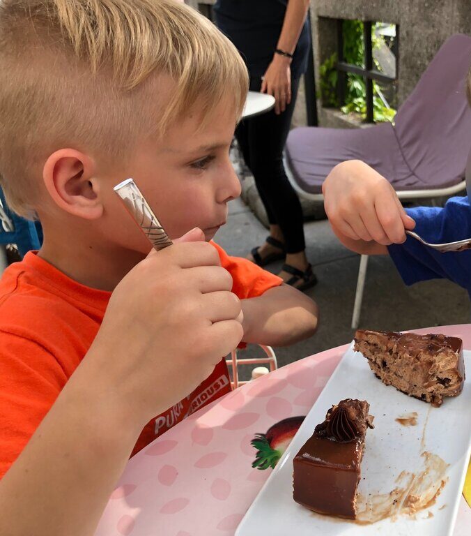 Child in an orange shirt is sitting at an outdoor cafe in Ljubljana holding a fork and looking at slices of chocolate cake on a white plate. One piece is topped with glossy chocolate glaze and a small swirl of cream, while another slice shows a creamy layered filling.