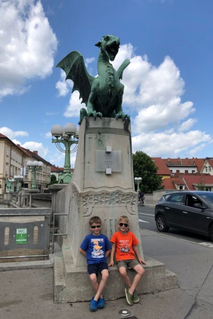 Two children sitting beneath the Ljubljana Dragon statue on Dragon Bridge. The large green dragon towers above them against a blue sky and red roofed buildings.