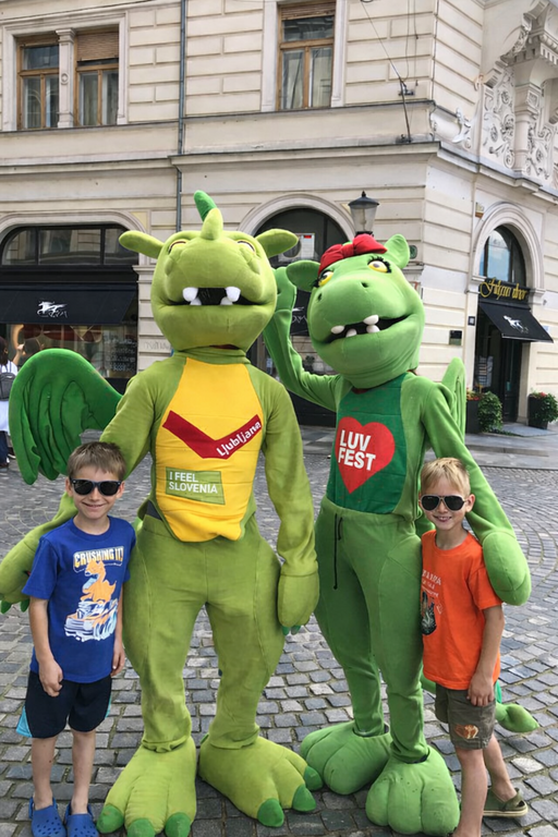 Two green dragon mascots wearing I Feel Slovenia and LUV FEST shirts pose with children on a cobblestone street in Ljubljana. The dragons represent the famous Ljubljana Dragon, the city symbol.