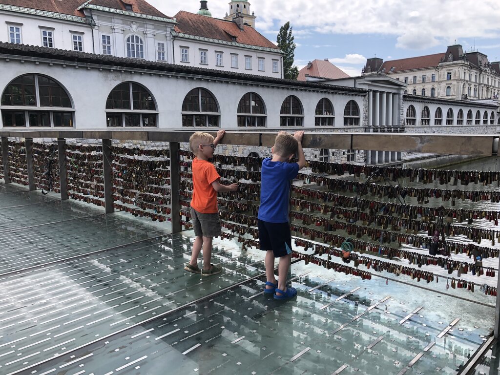 Two young boys stand on a glass walkway covered in love locks overlooking the Ljubljanica River with historic buildings in the background in Ljubljana Slovenia.
