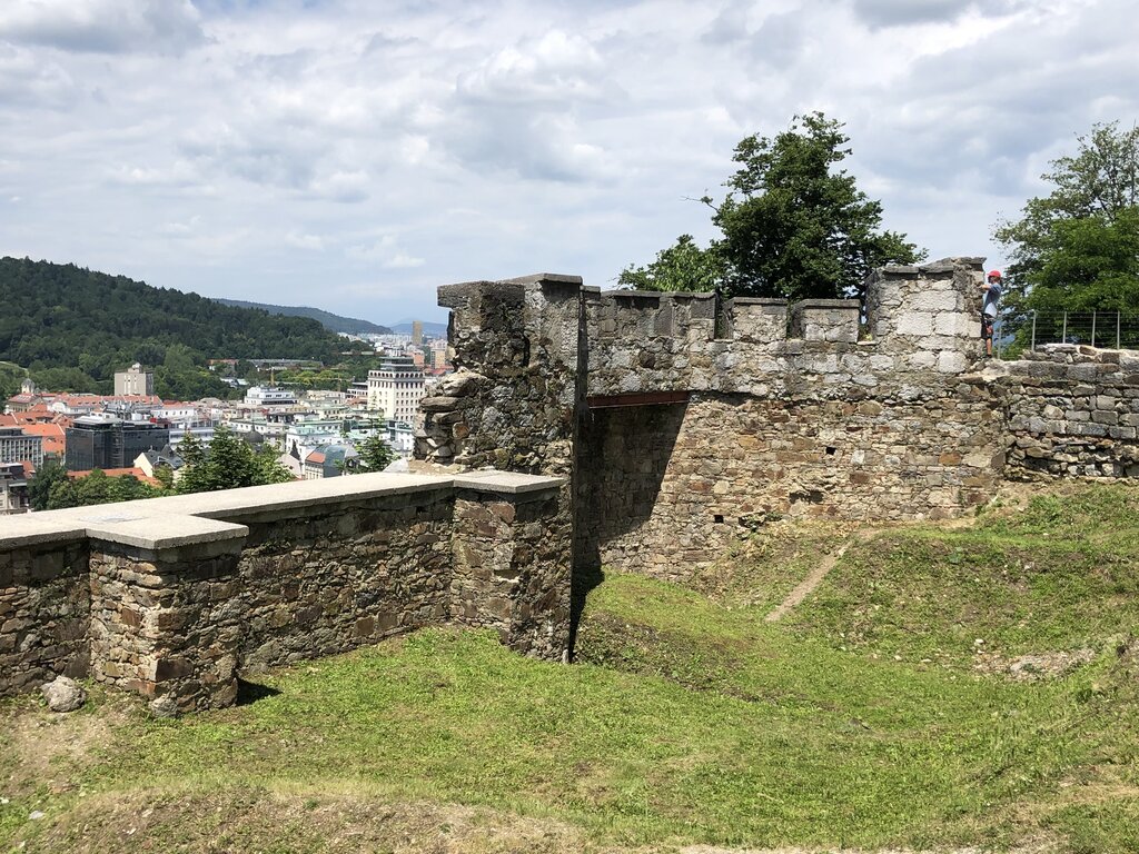 Stone walls and grassy courtyard inside Ljubljana Castle overlooking the city below. The medieval fortifications frame views of rooftops and distant hills.