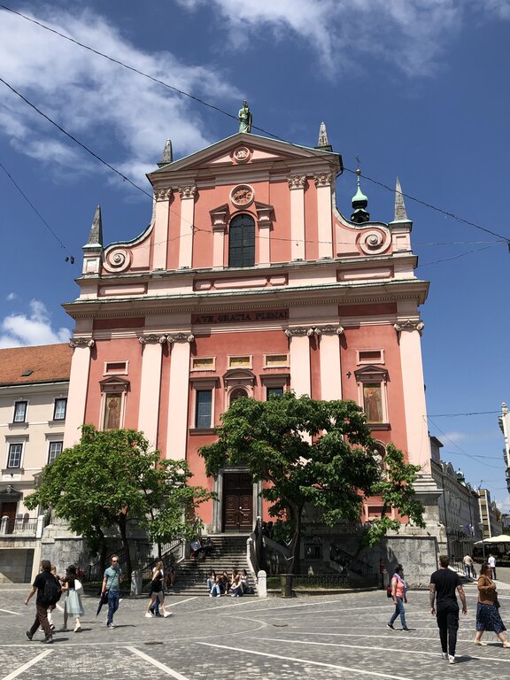 Front view of the pink Franciscan Church in Prešeren Square with people walking across the cobblestones. Decorative columns and statues stand out against a bright blue sky.