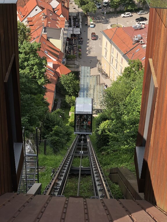 Ljubljana Castle funicular descending a steep green hillside toward the old town streets below. The glass cabin carries visitors up and down the hill for easy access to the castle.