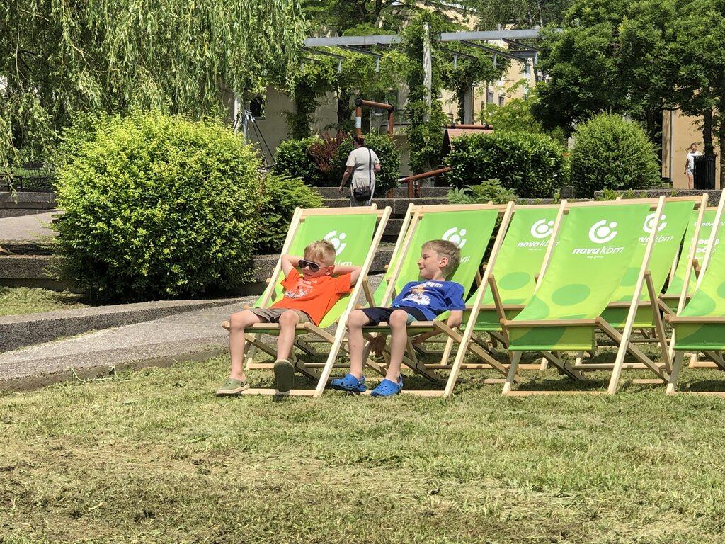 Two children relaxing in bright green deck chairs in a riverside park in Ljubljana. The chairs sit on grass surrounded by trees and garden paths.