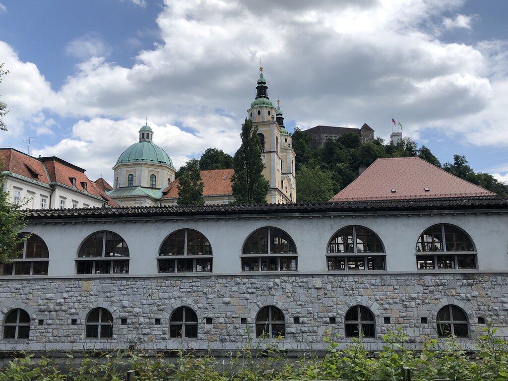 View of Ljubljana Cathedral domes and Ljubljana Castle on the hill behind historic riverside buildings. Arched windows of the market line the stone embankment along the Ljubljanica River.