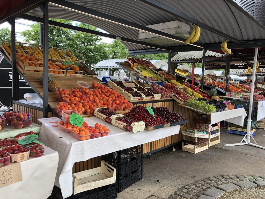 Colourful fruit stands at Ljubljana Central Market filled with cherries, apricots, strawberries, grapes, and bananas. Wooden crates and handwritten price signs create a lively open air market scene.