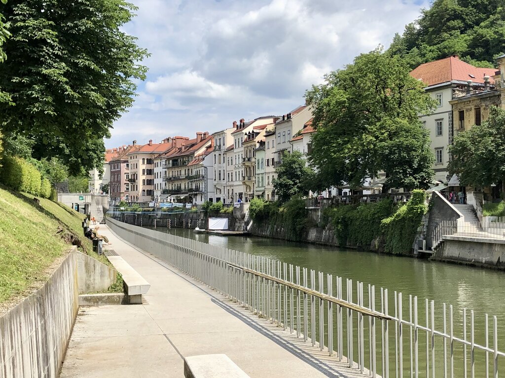 Riverside path in Ljubljana with historic buildings on one side and the Ljubljanica River on the other. A low railing separates the walkway from the water, making it easy to stroll safely. The flat pedestrian paths are ideal for exploring Ljubljana with kids.
