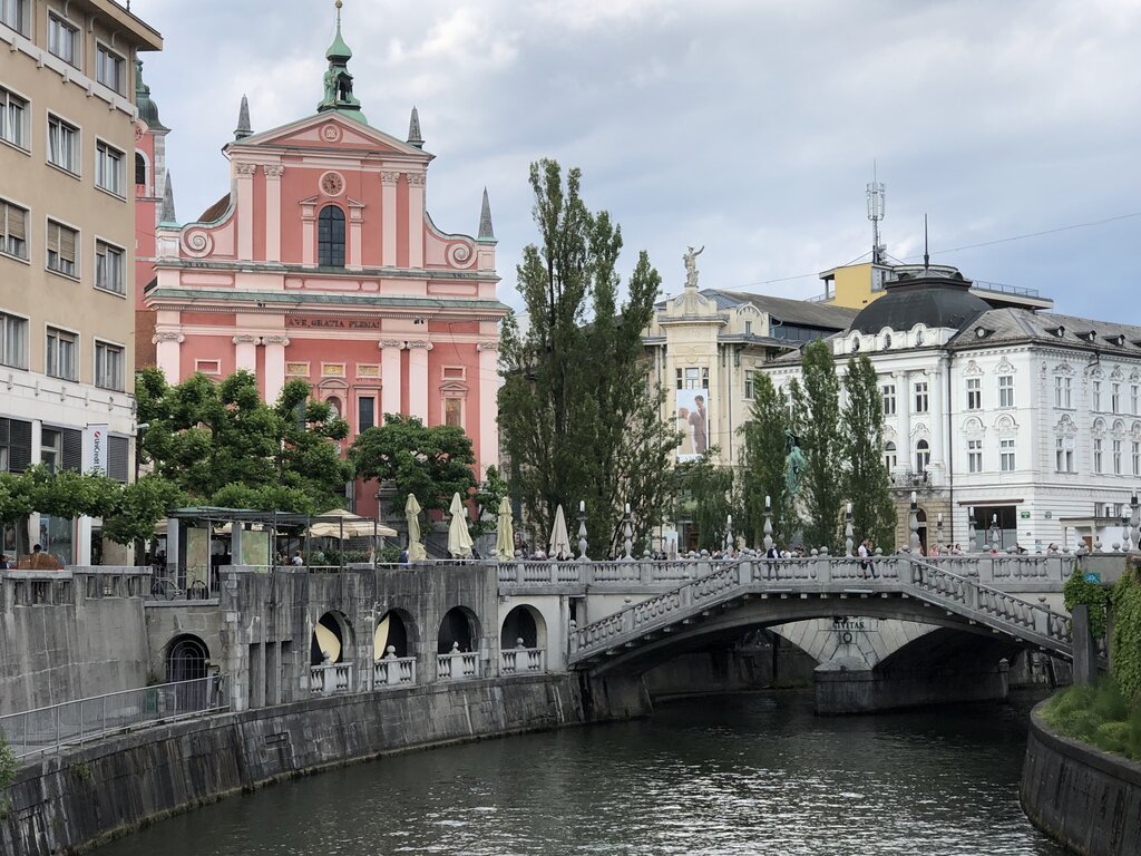 Triple Bridge in Ljubljana with the pink Franciscan Church rising behind it and the Ljubljanica River flowing below. Families and visitors stroll across the stone bridge lined with trees and riverside cafes. This central gathering spot is a must see when visiting Ljubljana with kids.