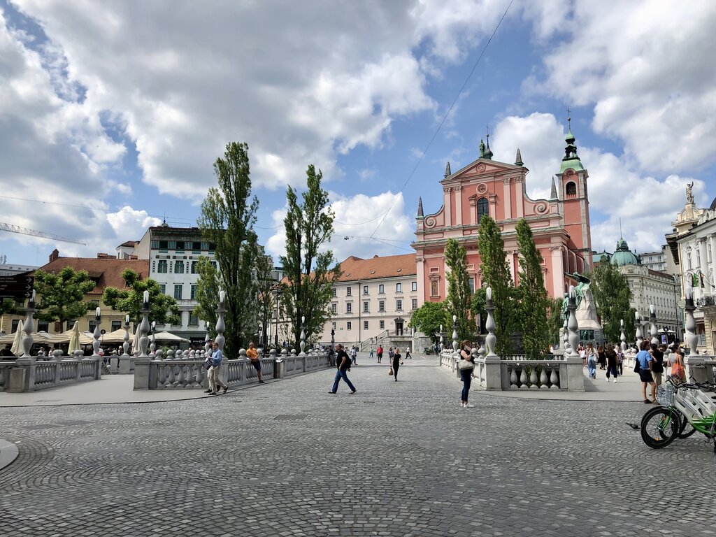 Wide view of Triple Bridge in Ljubljana filled with pedestrians under a bright blue sky. The pink church and historic buildings frame the cobblestone square.
